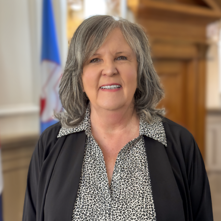 Portrait of Arna Hoffman, newly appointed Mesa County Treasurer, standing indoors with the Colorado state flag and Mesa County flag in the background.