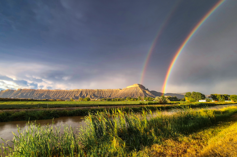 A vibrant double rainbow arches over Mount Garfield, with a serene landscape featuring lush green fields, tall grasses, and a reflective canal in the foreground.