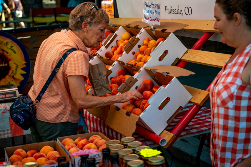 A woman examines fresh peaches displayed in cardboard boxes at a farmers' market booth. The stand features neatly arranged produce and jars of local goods.