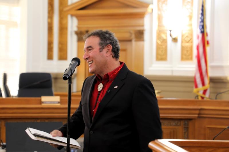 Man (Mesa County Commissioner JJ Fletcher) standing at microphone smiling and holding a notebook. 