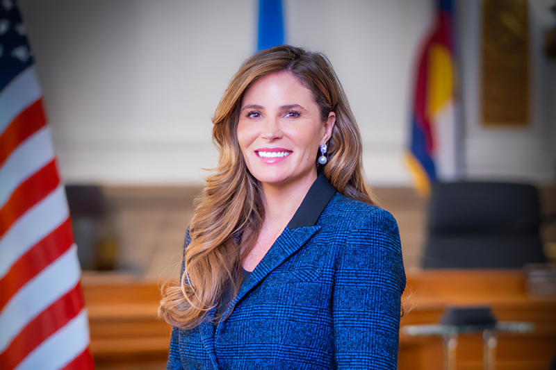 A professional portrait of a woman with long, wavy hair wearing a blue blazer, smiling in a courtroom or government setting. An American flag and other banners are visible in the background.