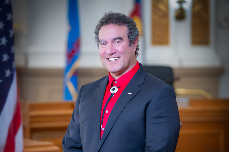 A professional portrait of a man with dark curly hair wearing a red shirt, black blazer, and bolo tie, smiling in a courtroom or government setting. An American flag and other banners are visible in the background.