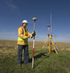 A surveyor in a yellow jacket and white hardhat uses survey equipment on a piece of undeveloped land.