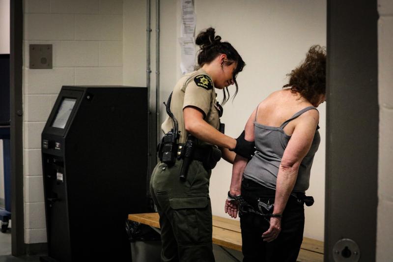 A young woman with dark hair in a tan uniform top with black pants an a utility belt pats down an older woman with fuzzy black hair. 
