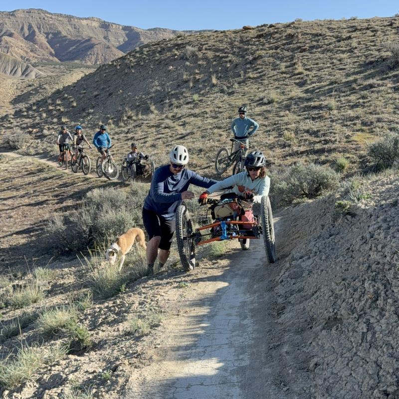 Seven people are biking a trail at North Fruita Desert along with two dogs. There are two people using specially modified electric motor assisted tricycles called adaptive mountain bikes. One person is guiding the adaptive biker through a narrow section of trail.