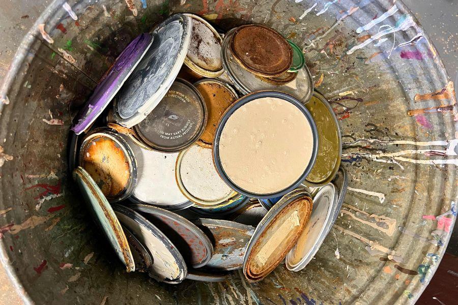 Aerial view of numerous paint can lids covered in leftover paint sitting in a metal trash can with leftover paint drippings. 