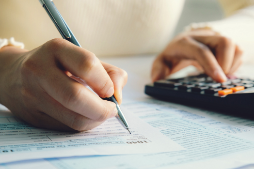 Close up image of hand holding a pen, filling out a tax form, as well as a hand inputting numbers on a calculator.