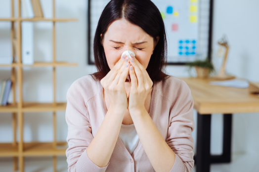 A woman with short, dark brown hair, is wearing a pink sweater and sneezing into a tissue.