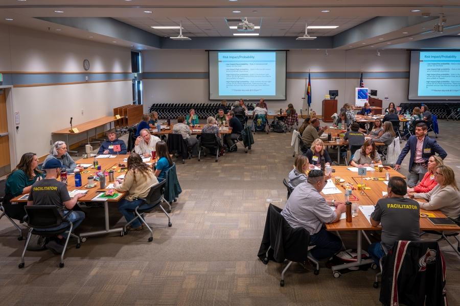 Groups of people sit at round tables in large room with projector screens. 