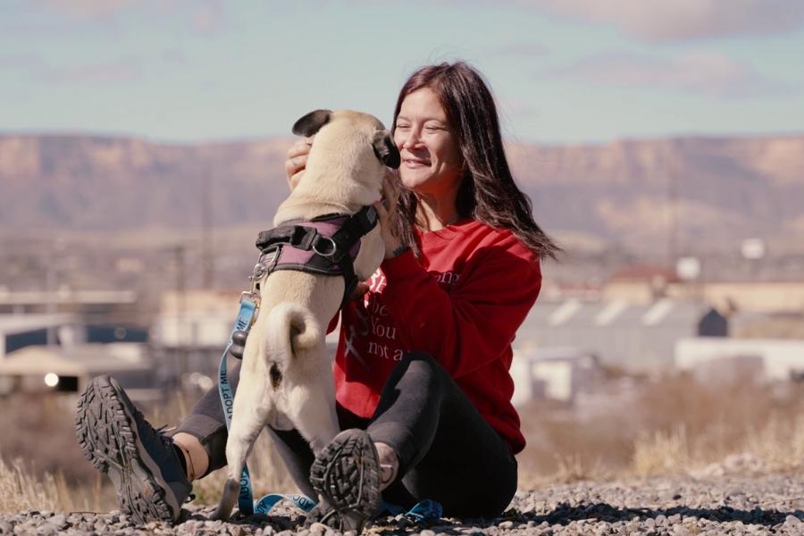 Woman smiling looking at Pug dog she is holding with mountain views behind her. 