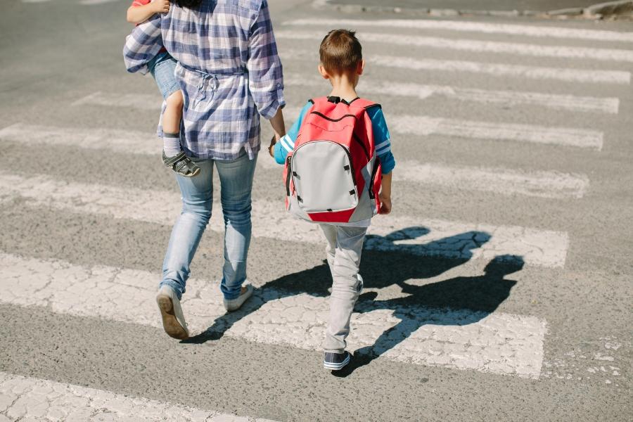 Woman holding child and holding hands with another child with red backpack on cross walk. 