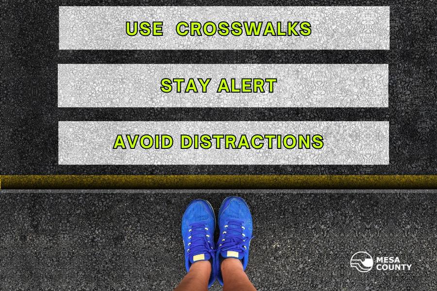 White crosswalk with green text reading, "USE CROSSWALKS STAY ALERT AVOID DISTRACTIONS: with a white Mesa County logo. 