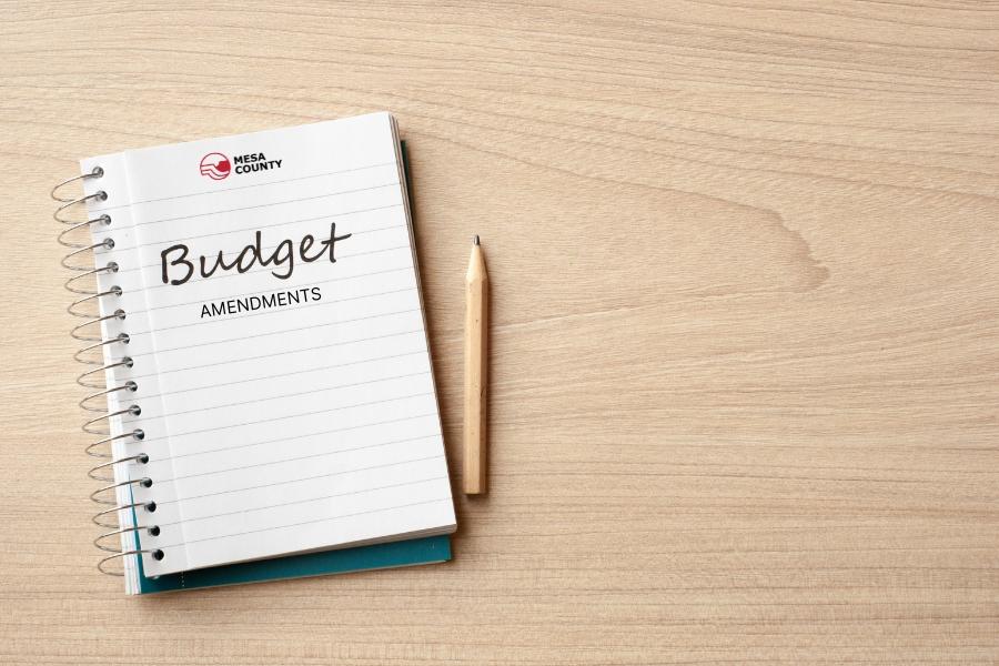 White notebook and pencil on light tan desk with black writing reading, "Budget AMENDMENTS," with the red and black Mesa County logo on the top.