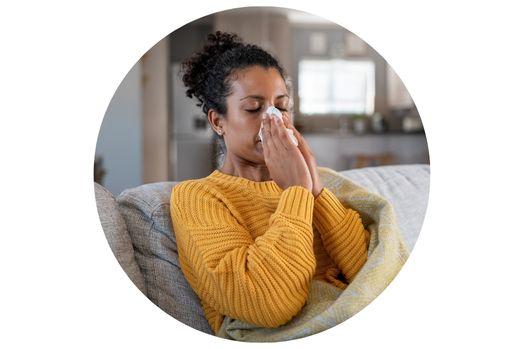 Woman wearing a gold sweater with black, curly hair pulled up into a pony tail, wrapped in a light yellow blanket, sitting on a gray couch. She is holding a white tissue up to her nose.