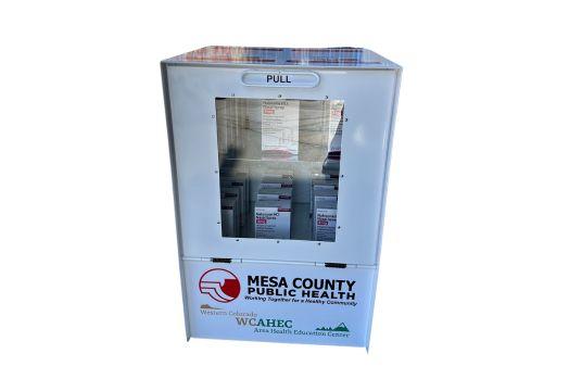 An image of a large, white, metal kiosk with a window below a handle that you pull down to open. On the kiosk are logos for Mesa County Public Health and Western Colorado Area Education Center.