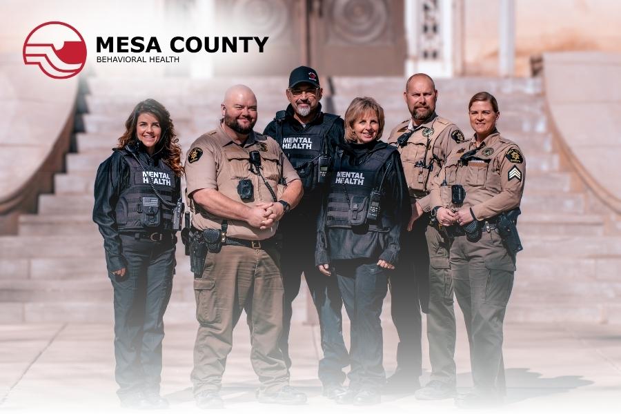 Group of law enforcement and mental health professionals smiling with the red Mesa County Behavioral Health logo and black text reading, "what do we do?" 