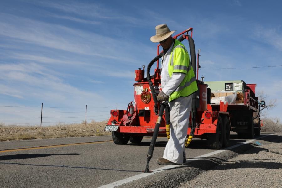 Man in bright yellow construction vest holding tool attached to large red truck that is dispersing black hot asphalt onto road. 