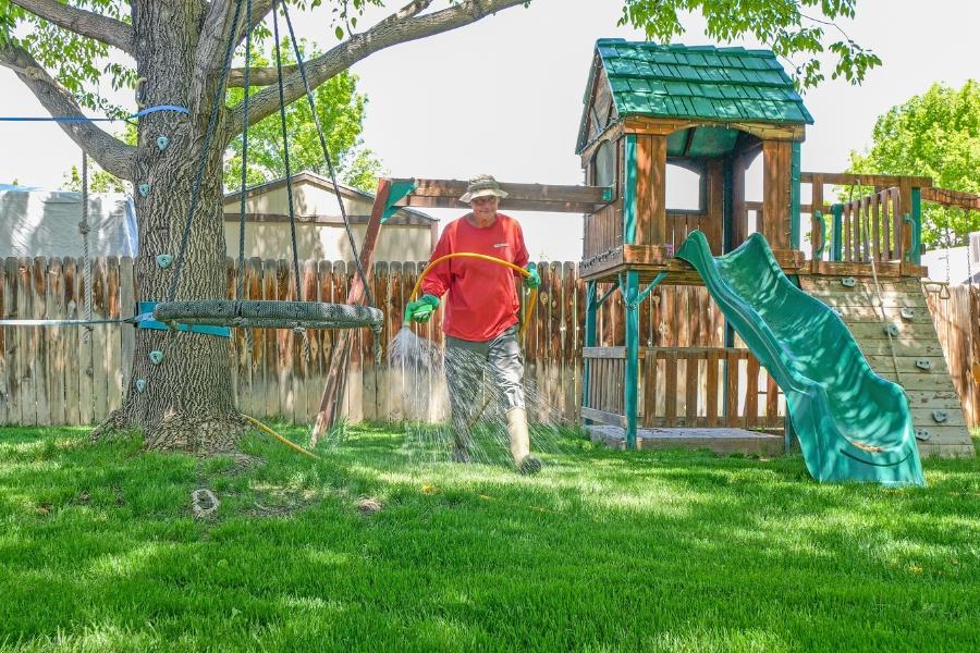 A man in a red shirt spraying grass with insecticides near a playground in a backyard. 