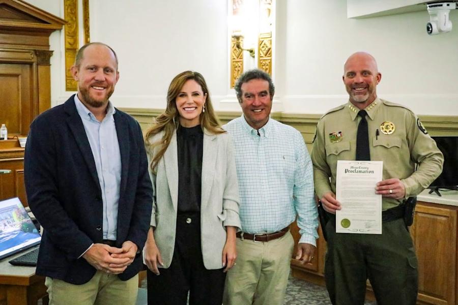 Four individuals are smiling and one in a Sheriff's Office uniform is holding a proclamation. 