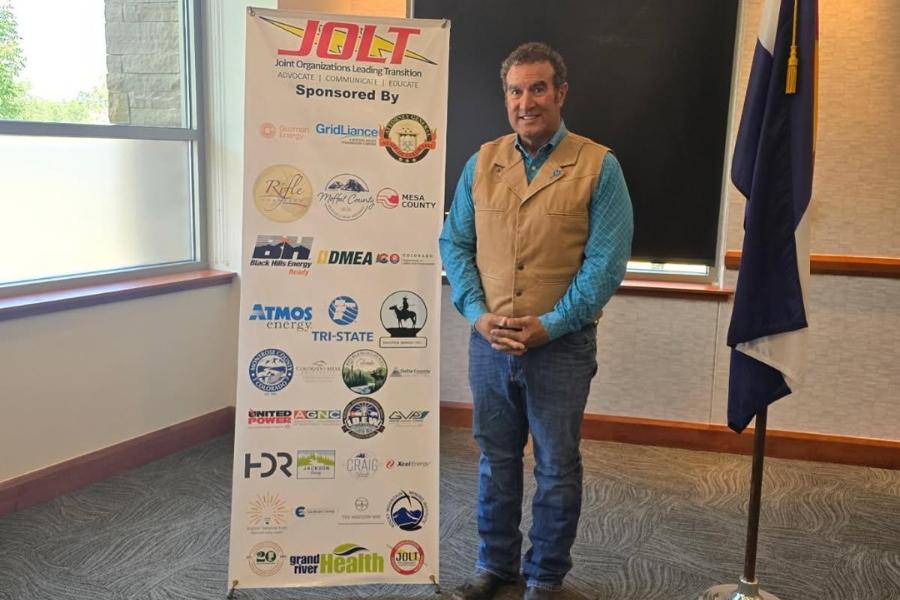 A man (JJ Fletcher, a Mesa County Commissioner), standing next to a banner that says, JOLT.