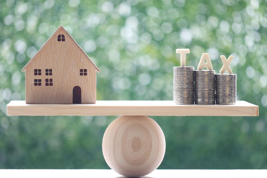 A toy house on a wooden scale next to change with letters reading tax above it.