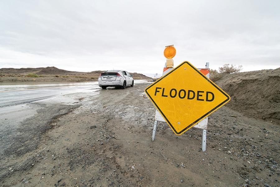 A car driving on a muddy road with a yellow sign reading, FLOODED. 