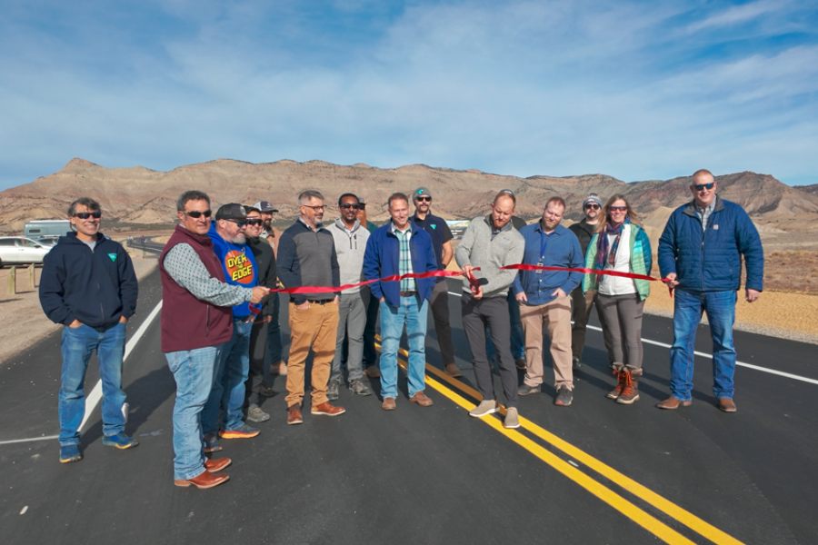 A group of people stand across a newly paved roadway holding a red ribbon during a ribbon-cutting ceremony, with desert hills and mountains in the background under a clear blue sky.