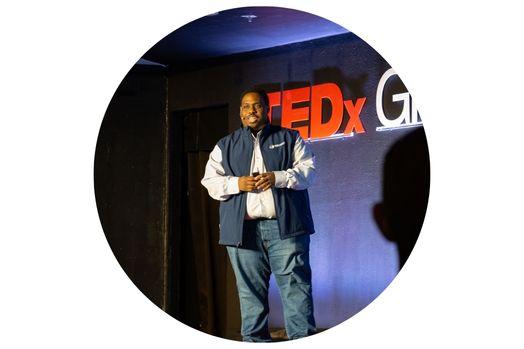 A person wearing jeans, a white button down shirt, and blue vest standing on stage at a tedx event wearing a microphone.