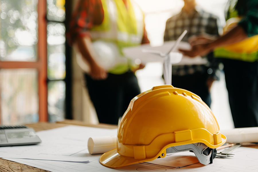 A yellow hard hat rests on construction plans spread across a table, with workers in safety vests discussing documents in the background.