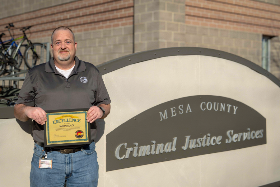 Jason Black, Mesa County Community Corrections case manager, stands outside the Mesa County Criminal Justice Services building holding a Certificate of Excellence.