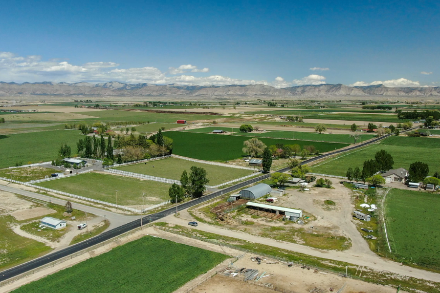 Aerial view of rural Mesa County showing farmland, irrigation fields and intersecting county roads, with homes and outbuildings in the foreground and mountains in the distance.
