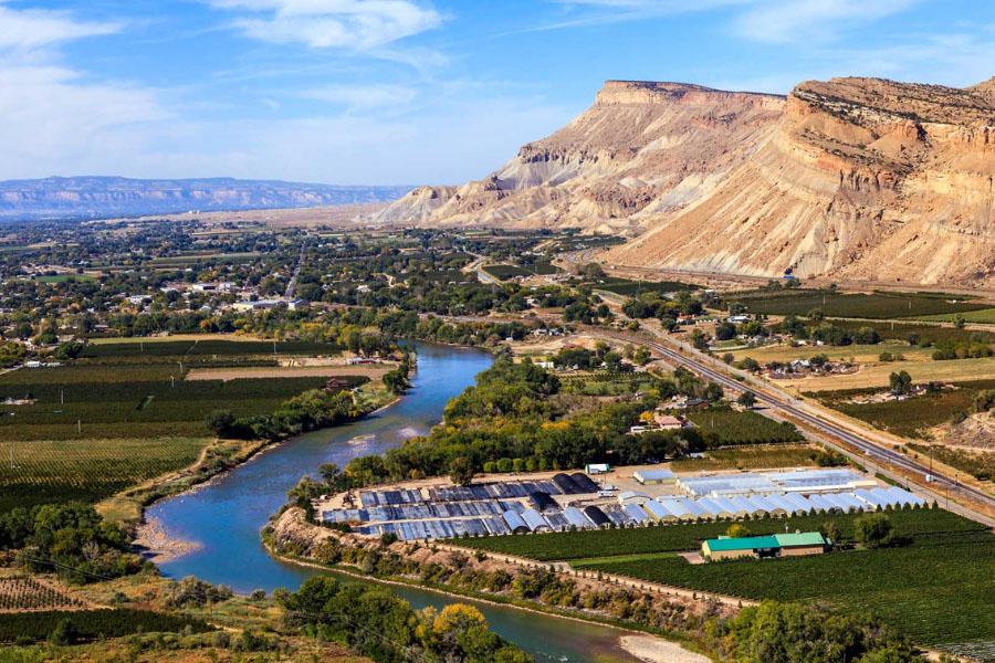 An aerial view of a river winding through a green agricultural valley with orchards and fields, a small town and roads nearby, and steep, layered red and tan mesas rising in the background under a blue sky.