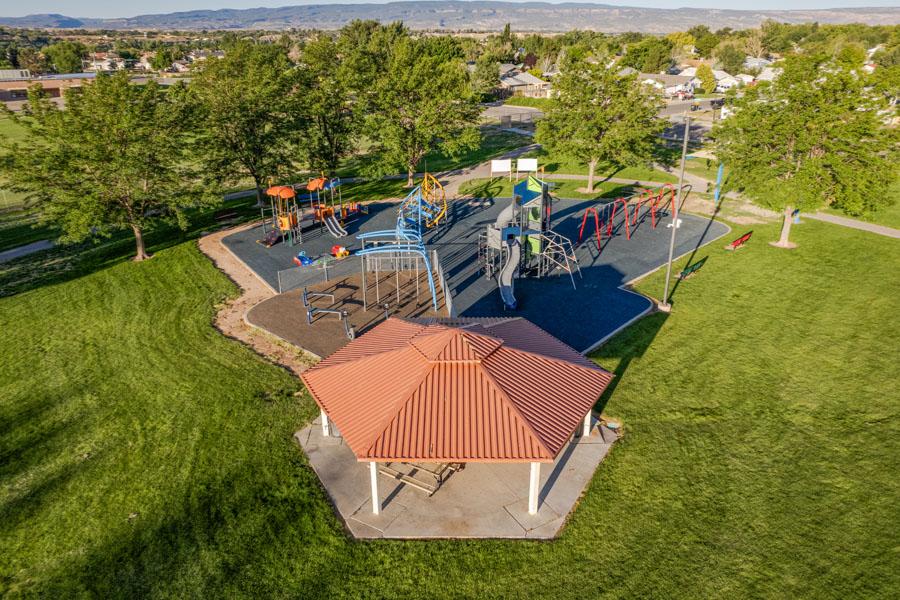 Aerial view of a Mesa County park featuring a large playground with slides, swings and climbing structures, surrounded by green grass and trees, with a covered picnic pavilion in the foreground and homes and mesas in the background.