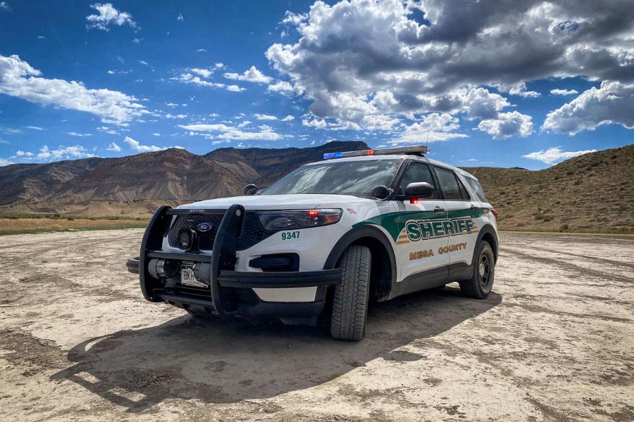 Mesa County Sheriff’s Office patrol vehicle parked on a dirt area with emergency lights on, set against desert terrain and a mountainous landscape under a partly cloudy sky.