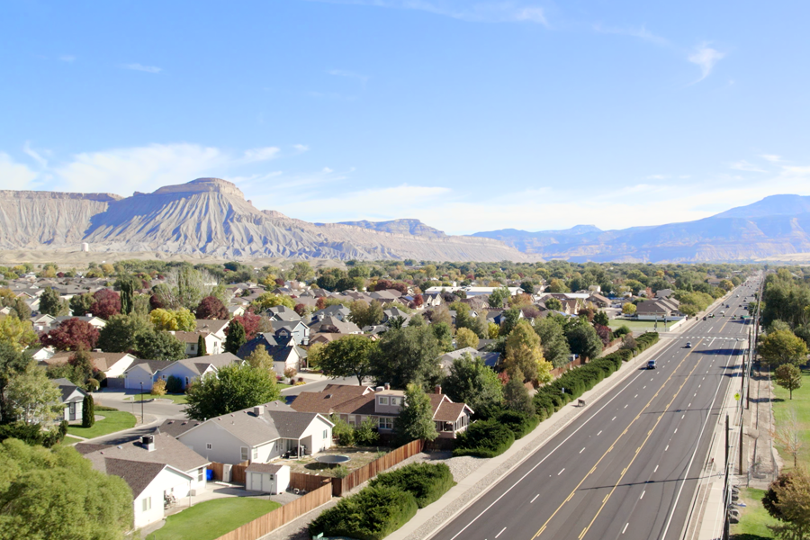 Aerial view of a residential neighborhood in the Fruitvale area of Mesa County with a multi-lane roadway running alongside homes, trees and sidewalks, under a clear blue sky.