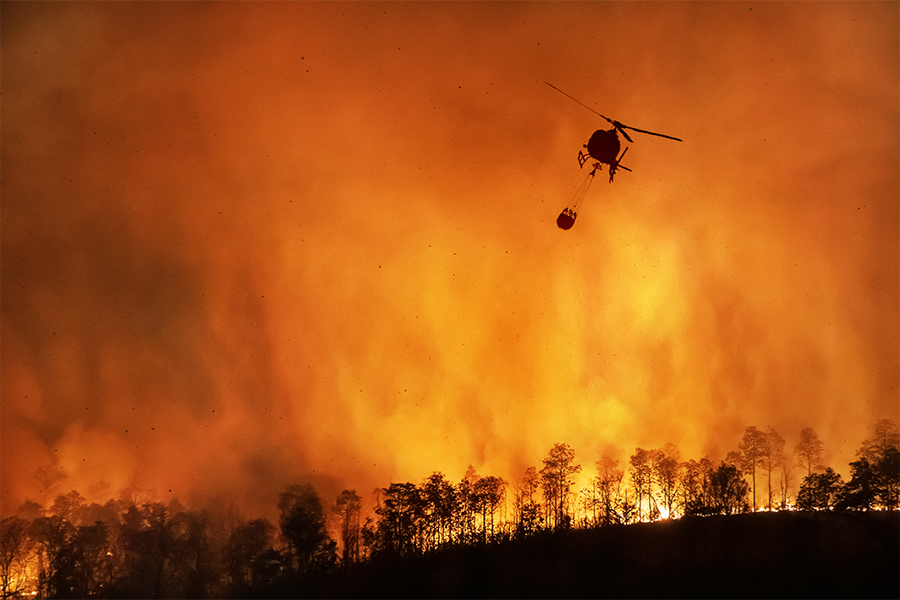 A helicopter drops water over an active wildfire as flames spread through a tree-covered hillside, with thick orange smoke filling the sky and silhouetting the aircraft and trees.