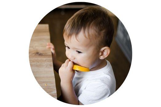 A toddler in a white shirt chews on a piece of orange while sitting at a wooden table.