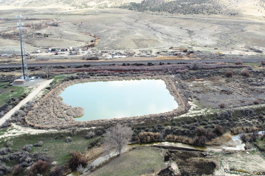 Aerial view of a wastewater lagoon near the Mack community in Mesa County, surrounded by dry hills, brush and rural land, with a roadway and utility tower nearby.