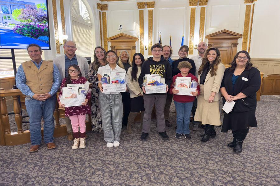 Four student winners stand at the front of the Mesa County Commissioners’ hearing room holding gift baskets and certificates, surrounded by county officials, school representatives and family members. The group poses for a photo beneath decorative columns and flags after the students were honored for their artwork in the “I Voted” sticker contest.
