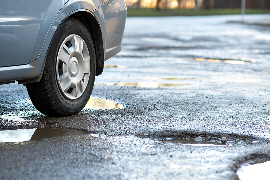 Car tire driving over a pothole on a worn road surface with standing water, highlighting road damage and maintenance needs.