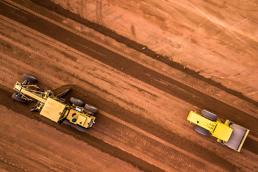 An aerial view of two road construction machines grading and preparing dirt roadway lanes at a construction site.