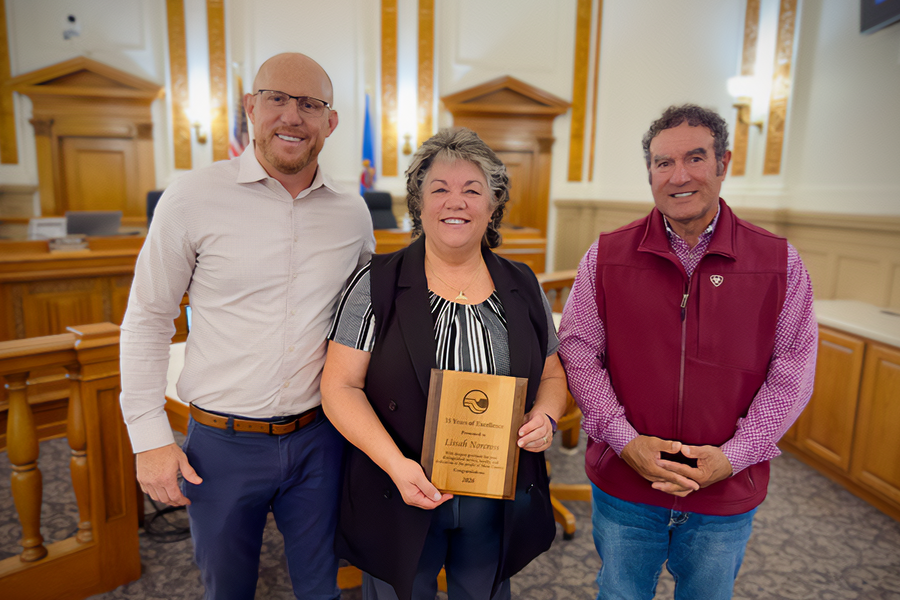 Mesa County Commissioner Cody Davis, Investigator Lissah Norcross and Mesa County Commissioner JJ Fletcher pose for a photo in the Mesa County public hearing room. Norcross holds a plaque recognizing her 35 years of service with the Mesa County Sheriff's Office as she prepares to retire.