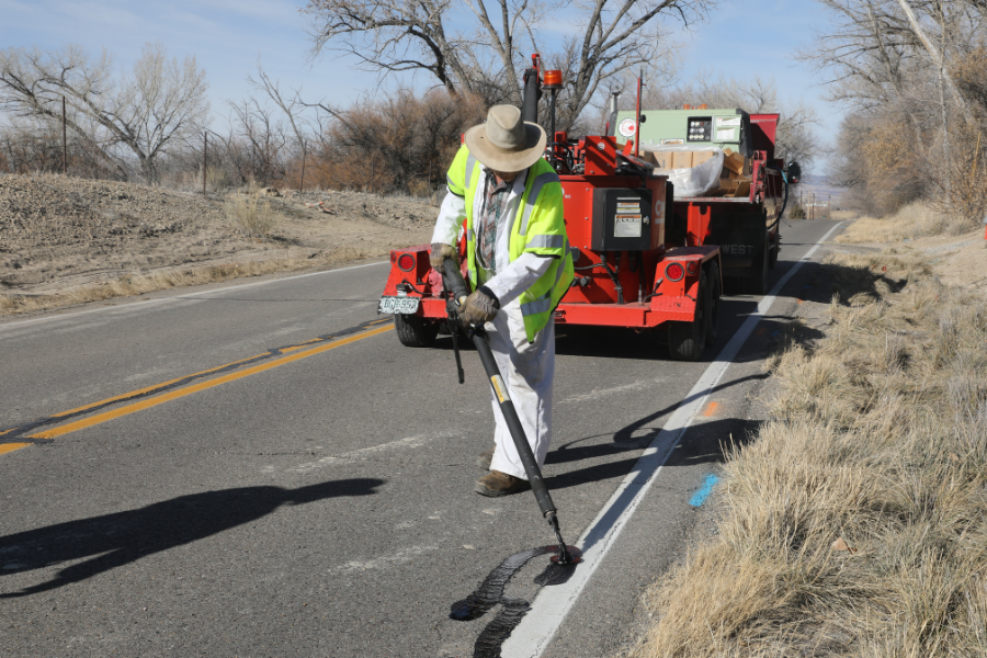 A Mesa County Road and Bridge worker applies crack seal to a paved rural road using specialized equipment, with a maintenance truck parked behind him.